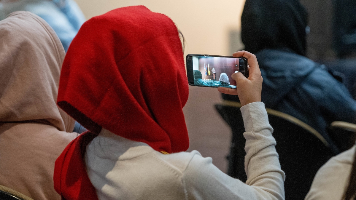 A woman in a headscarf films a speaker on her phone