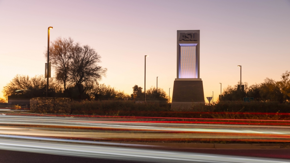 An ASU sign stands silhouetted against a sunset as traffic lights zoom by in the foreground