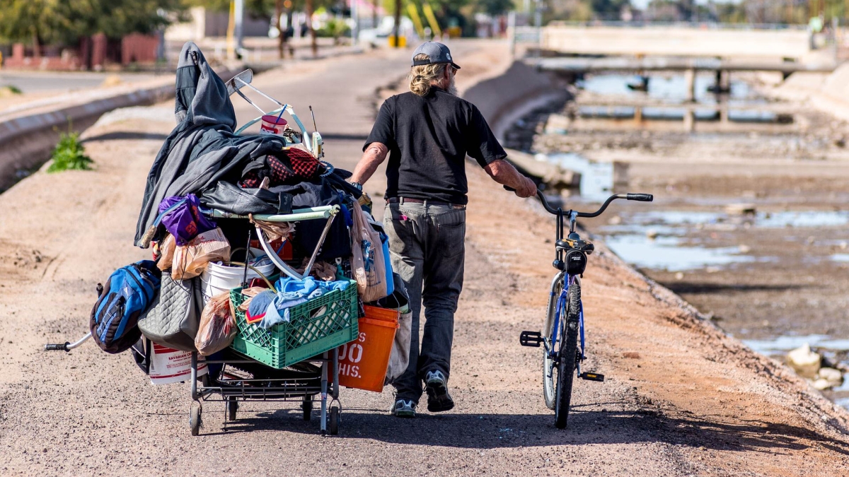 A homeless man walks along a Phoenix canal with a cart holding his possessions and a bicycle.