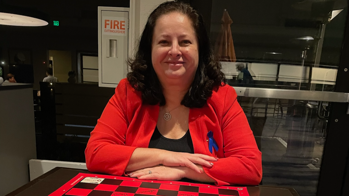Anjelina Belakovskaia sitting in front of a red and black chess board smiling at the camera.