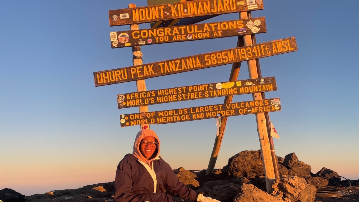 Lauren Crenshaw on Mt. Kilimanjaro sitting in front of a sign reading "Mount Kilimanjaro" and including other details about her exact location.