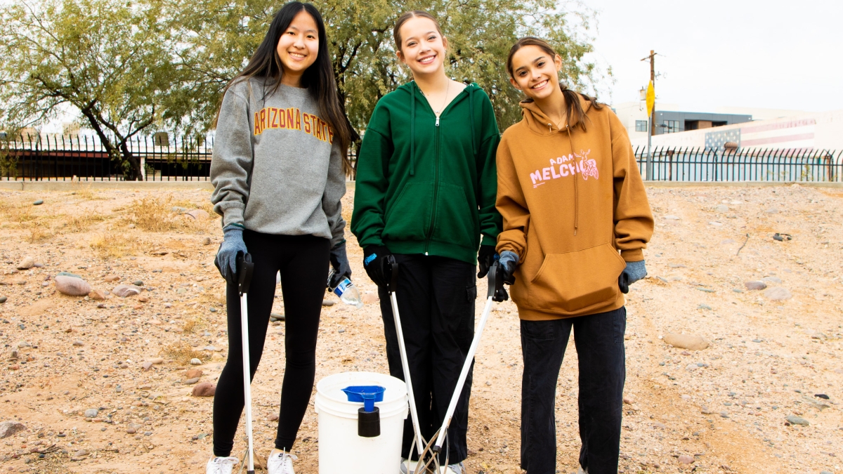 Students pose for a group photo holding trash pickers by a bucket in an outdoor setting.