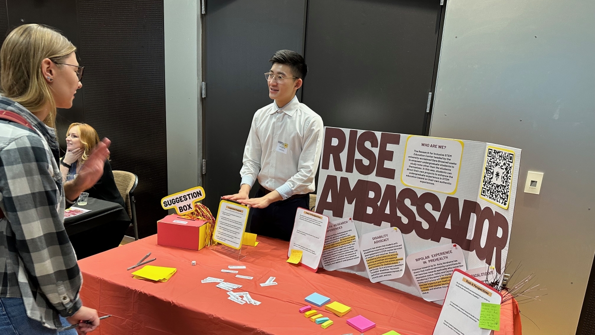 Photo of two students talking at a table with a poster that reads "RISE Ambassador."