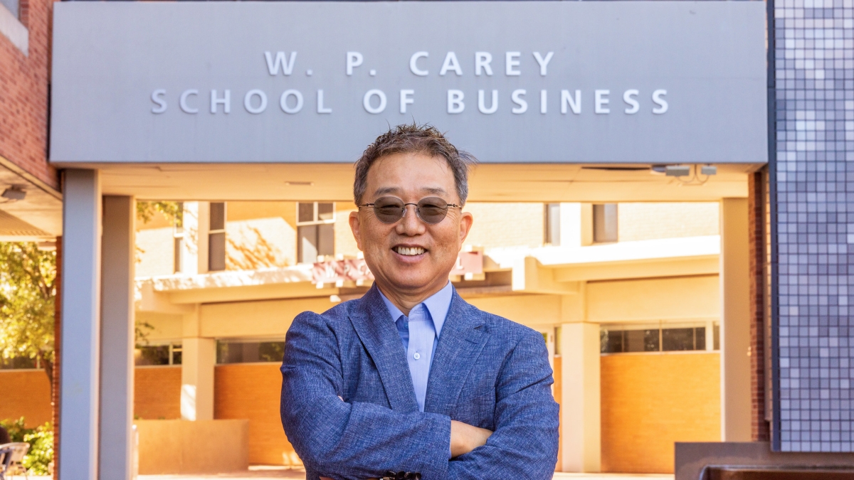 Thomas Choi in sunglasses, smiling with arms folded, standing in front of building reading "W. P. Carey School of Business."