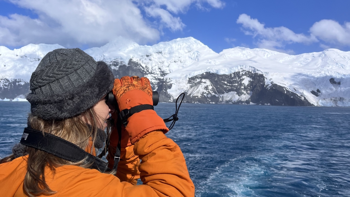 A woman in an orange jacket looking through binoculars at the Antarctic ocean.