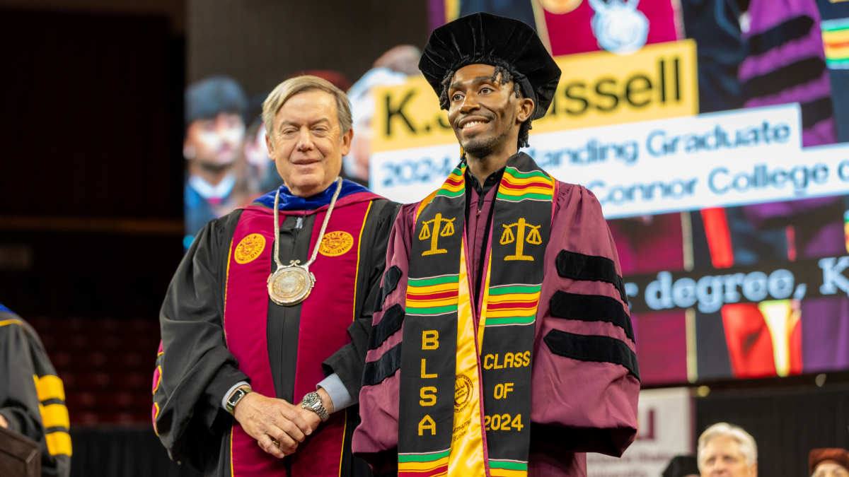 A law graduate in commencement regalia smiles from the stage as ASU President Crow stands to the side