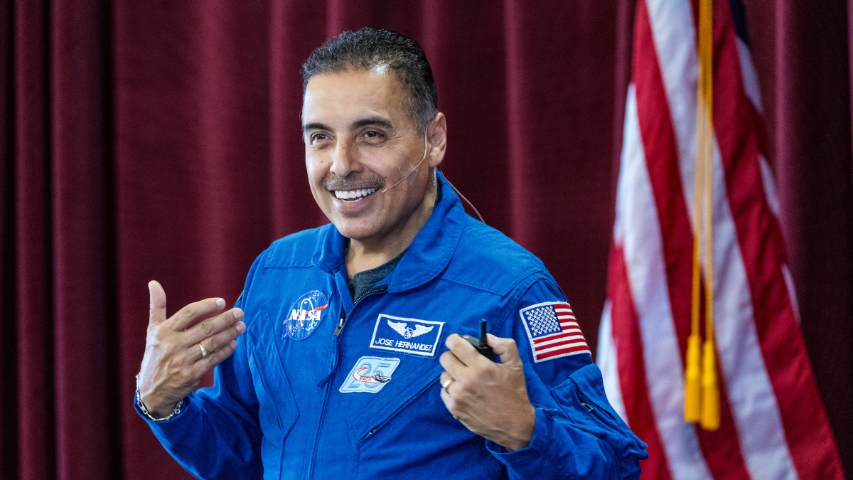 Man wearing a NASA flight suit stands in front of an American flag as he speaks to an unseen audience.