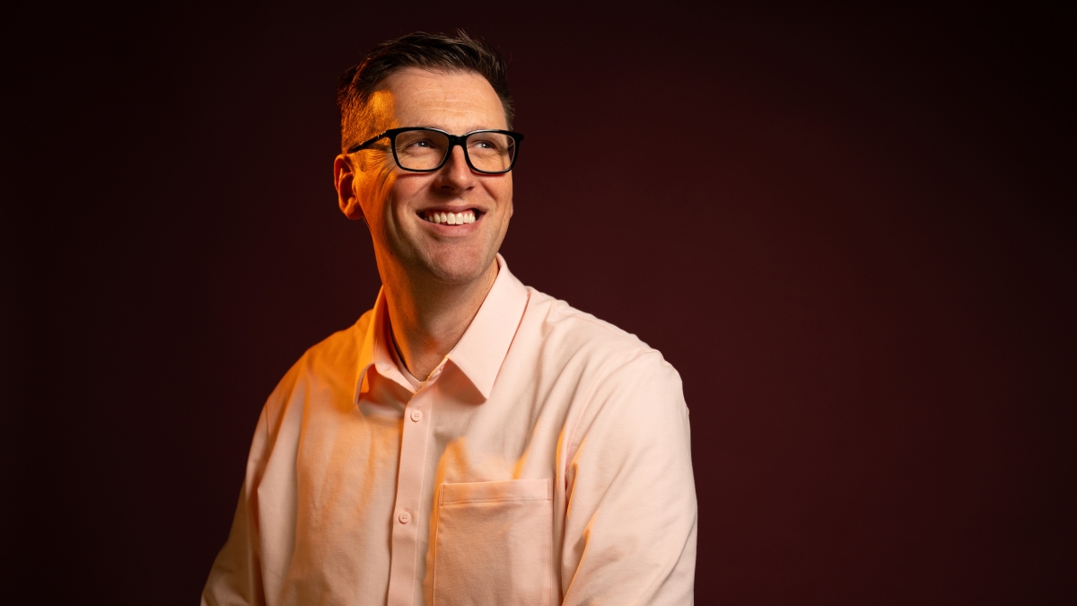 A studio portrait of Kyle Jensen, wearing a white shirt on a dark background lit with orange lighting