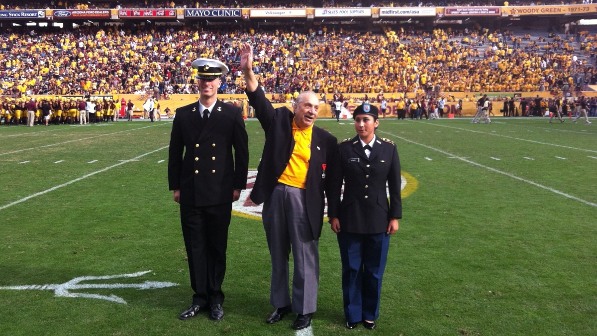 Elderly man waving to a crowd as he stands on a football field next to two servicemembers at Mountain America Stadium in Tempe, Arizona.