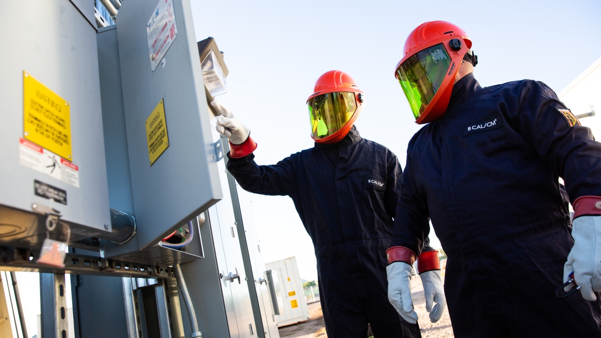 Two researchers wearing orange face guards and blue jumpsuits check an electrical panel.