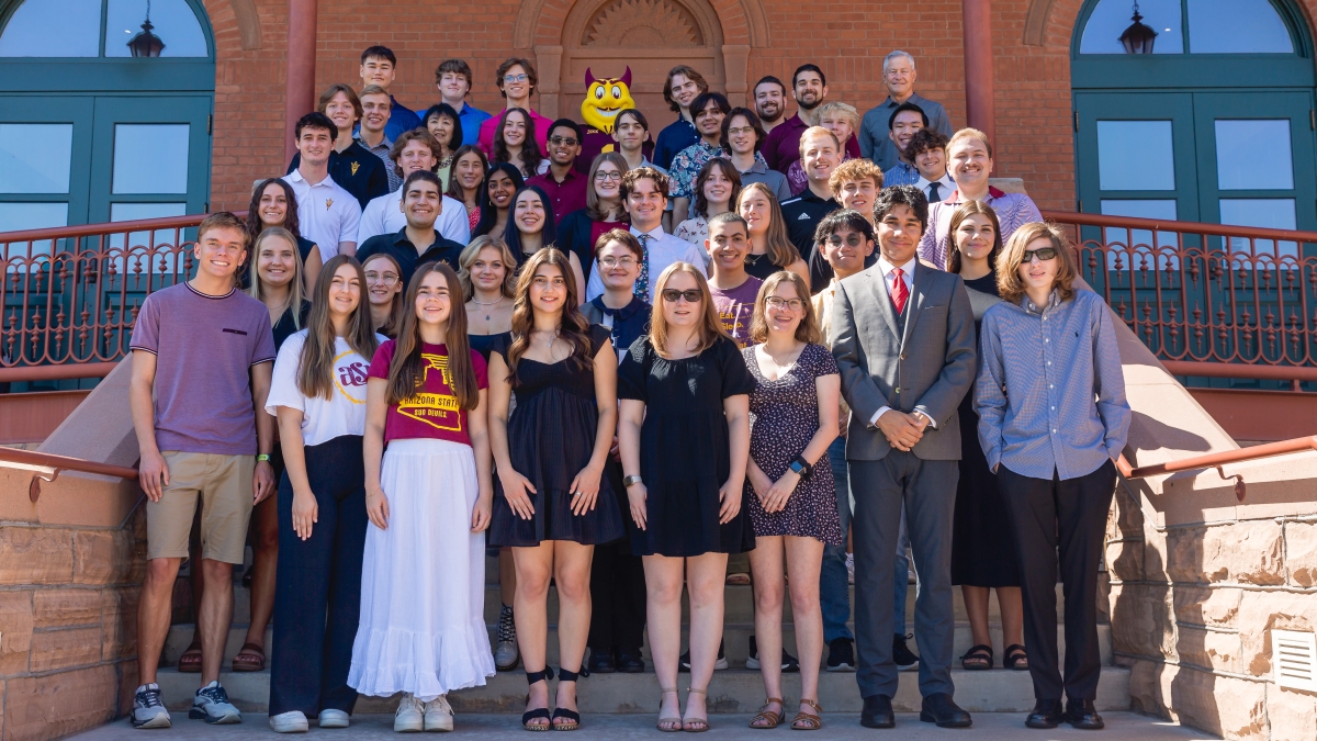 A large group of people posing on the steps of a red brick building with arched doors. ASU's mascot "Sparky" stands near the center.