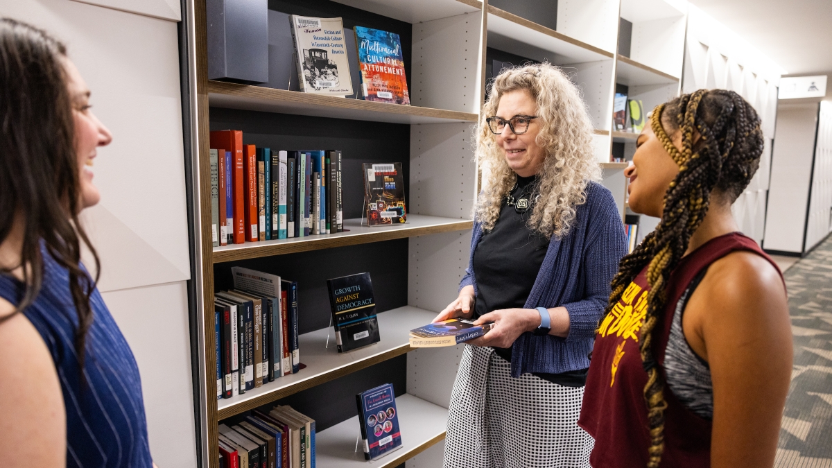 Three people interacting next to shelves of books in library