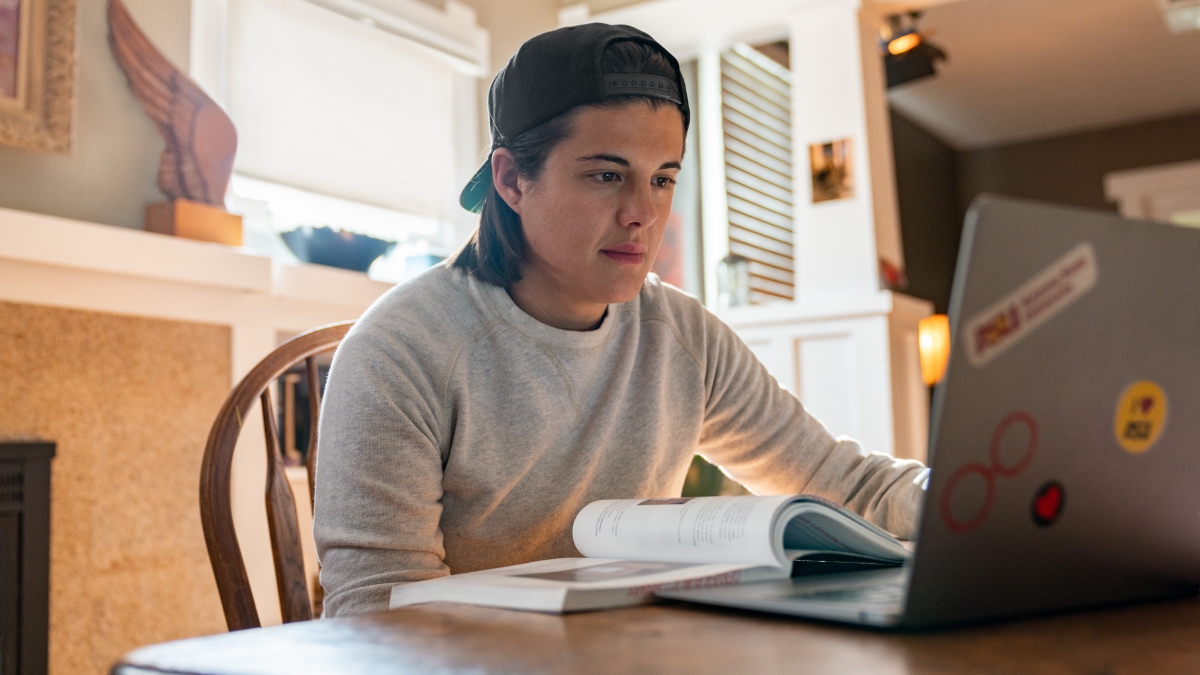 Person wearing a black backward cap and grey sweater, seated at a wooden table with a laptop and an open book.