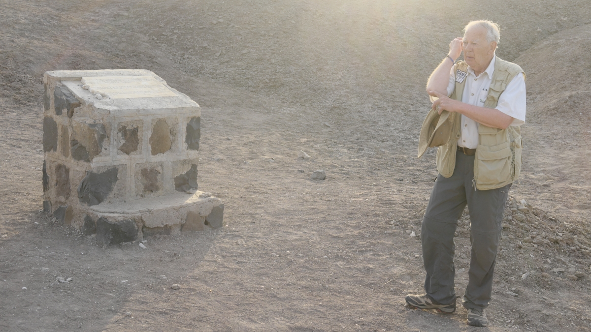 An older man points to his elbow while standing near a monument plaque in a barren dirt field