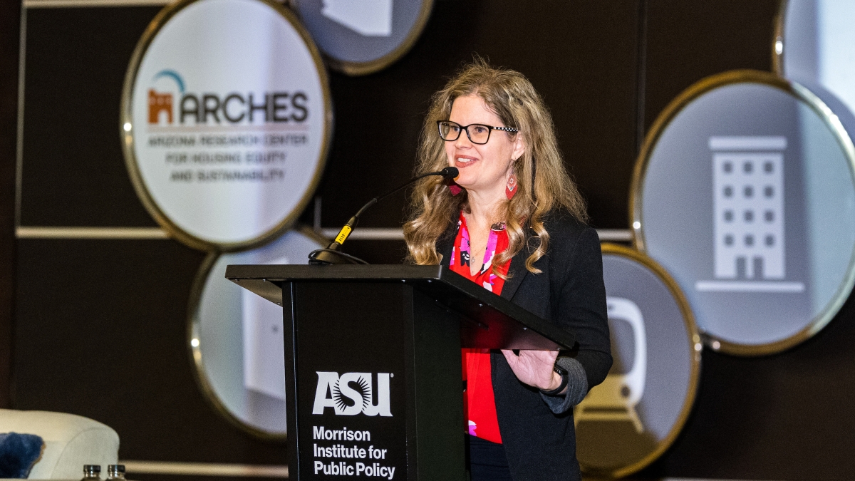 A woman with glasses stands behind a lectern smiling.