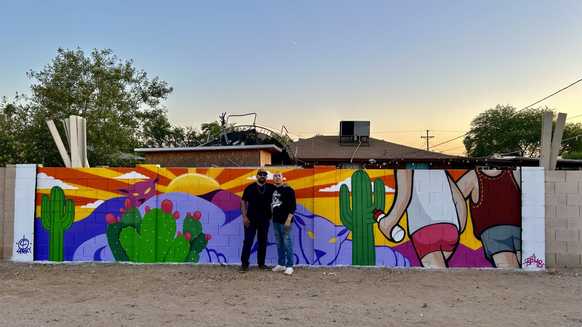 Two men stand in front of a mural of people hiking in a desert landscape.