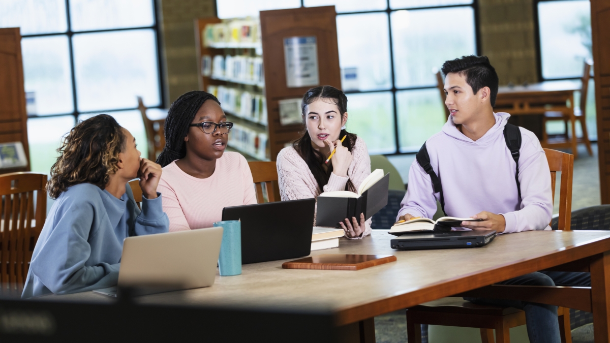 Group of teens sitting around a table in a library talking.