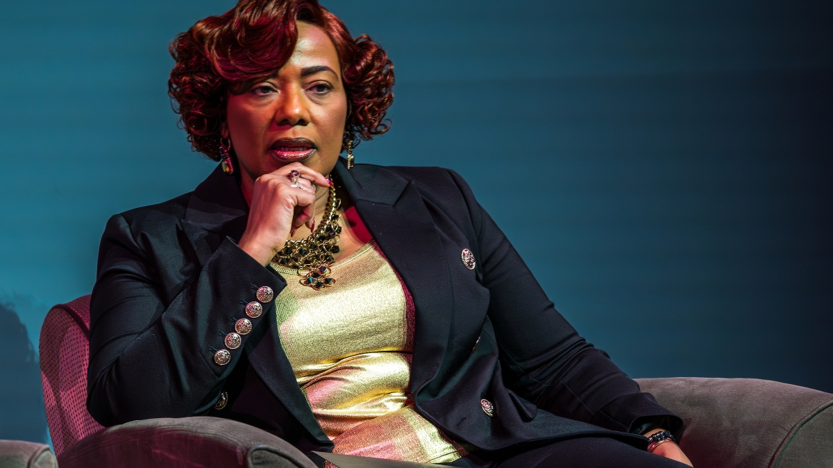 Black woman in black blazer and gold blouse sits in chair on stage during talk