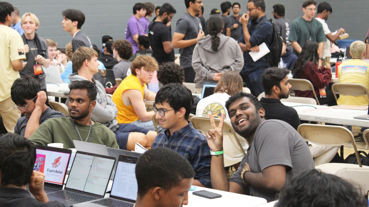 A large group of students sit at tables with laptops and mingle in a large room 