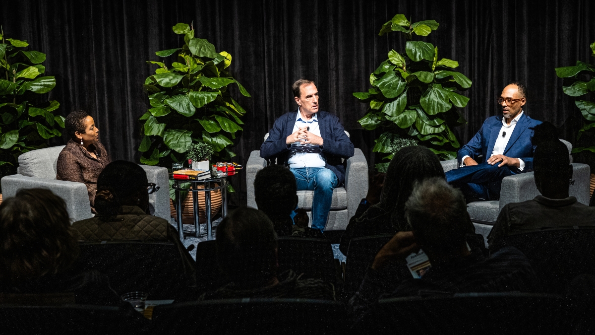 Three people sitting on stage in oversize gray chairs talking to audience with green plants behind them