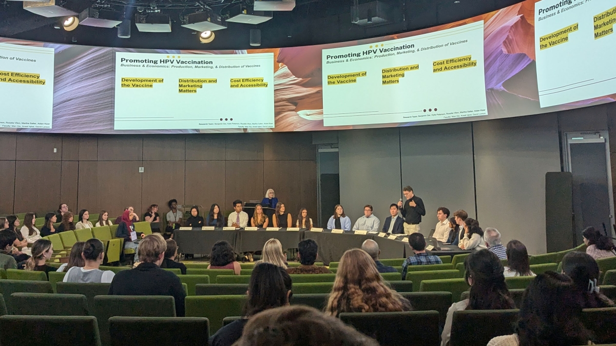 A lecture hall where a panel of students speaks to an audience with a series of screens above them.