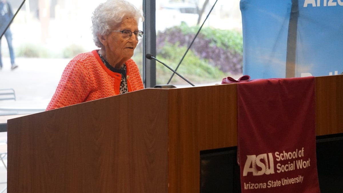 Elderly woman stands behind a podium while speaking into a microphone.