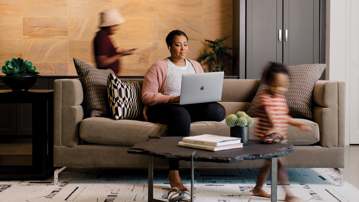 A woman works on her laptop while seated on a couch as her two children move around