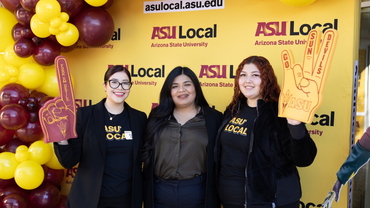 Three women posing for photo, with two on outside holding foam fingers, in front of ASU Local background.