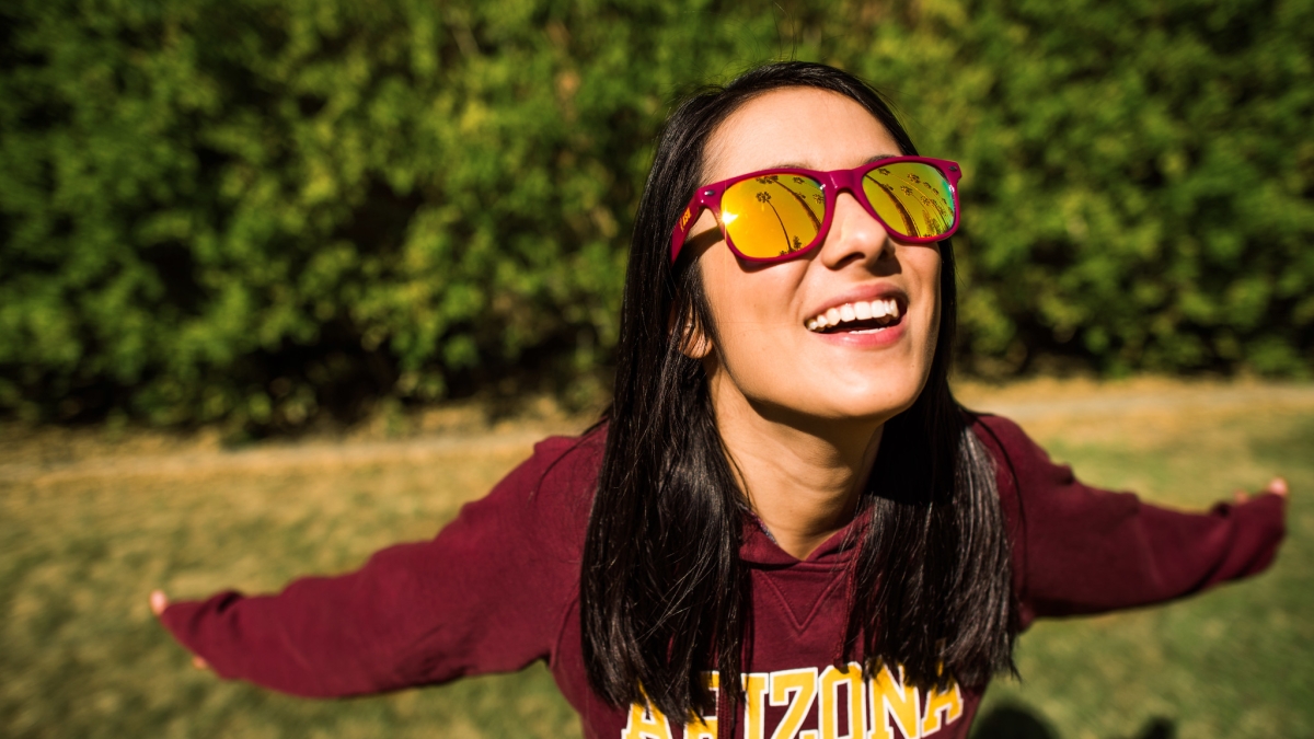 A women stands on a lawn with an ASU sweatshirt and matching sunglasses