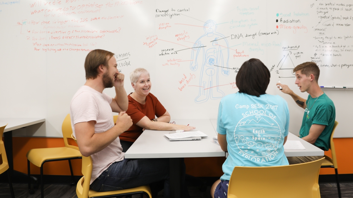 Students gathered at a table in front of a whiteboard in a classroom.