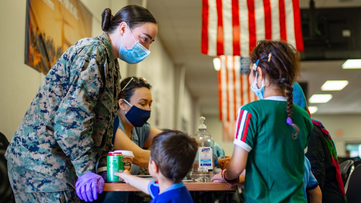 Helen Murray in military fatigues wearing a mask and standing next to children near a table with U.S. flags hanging in the background.