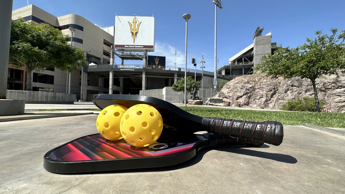 Gold whiffle balls stacked between two paddles rest on the cement in front of Sun Devil Stadium entrance with a pitchfork sign in background