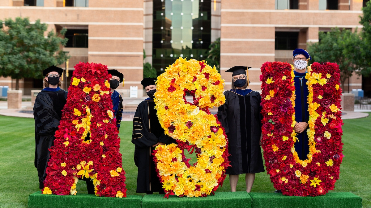 ASU West faculty members in commencement regalia and masks pose next to a flower arrangement that spells out 'ASU' on Fletcher Lawn