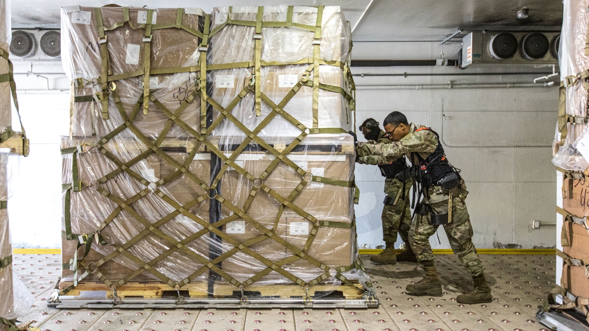 Two airmen wearing an ASU exoskeleton to help with movement push a pallet piled high with supply boxes