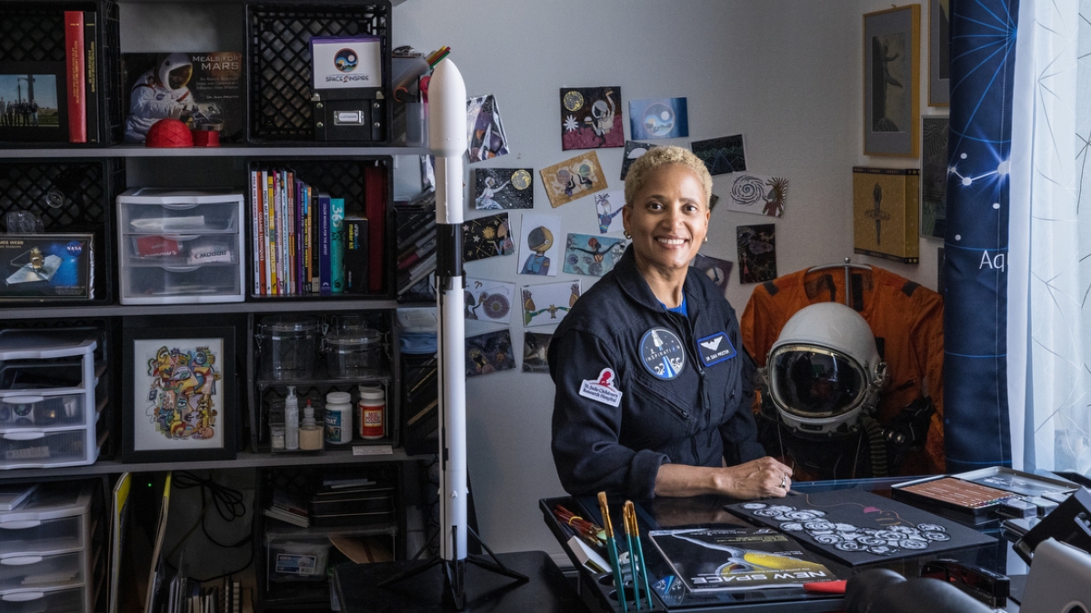 Smiling woman in a NASA space suit sitting in an office surrounded by space-themed paraphernalia.