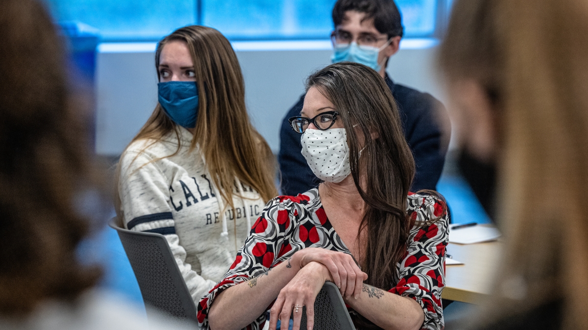 Woman seated in a classroom, wearing a face mask, looking back over her chair toward a student.