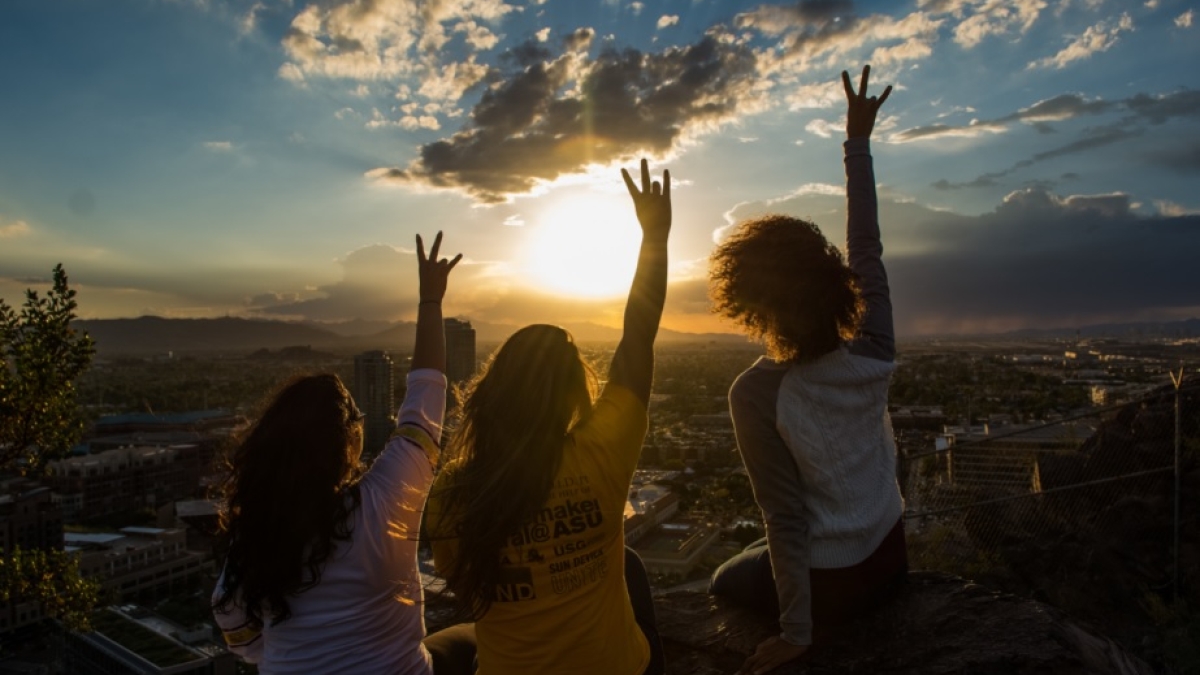 three women pictured from behind as they raise their hands toward a sunset
