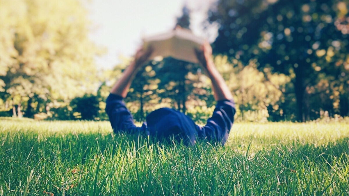 person holding and reading a book while they lie in the grass on a sunny day