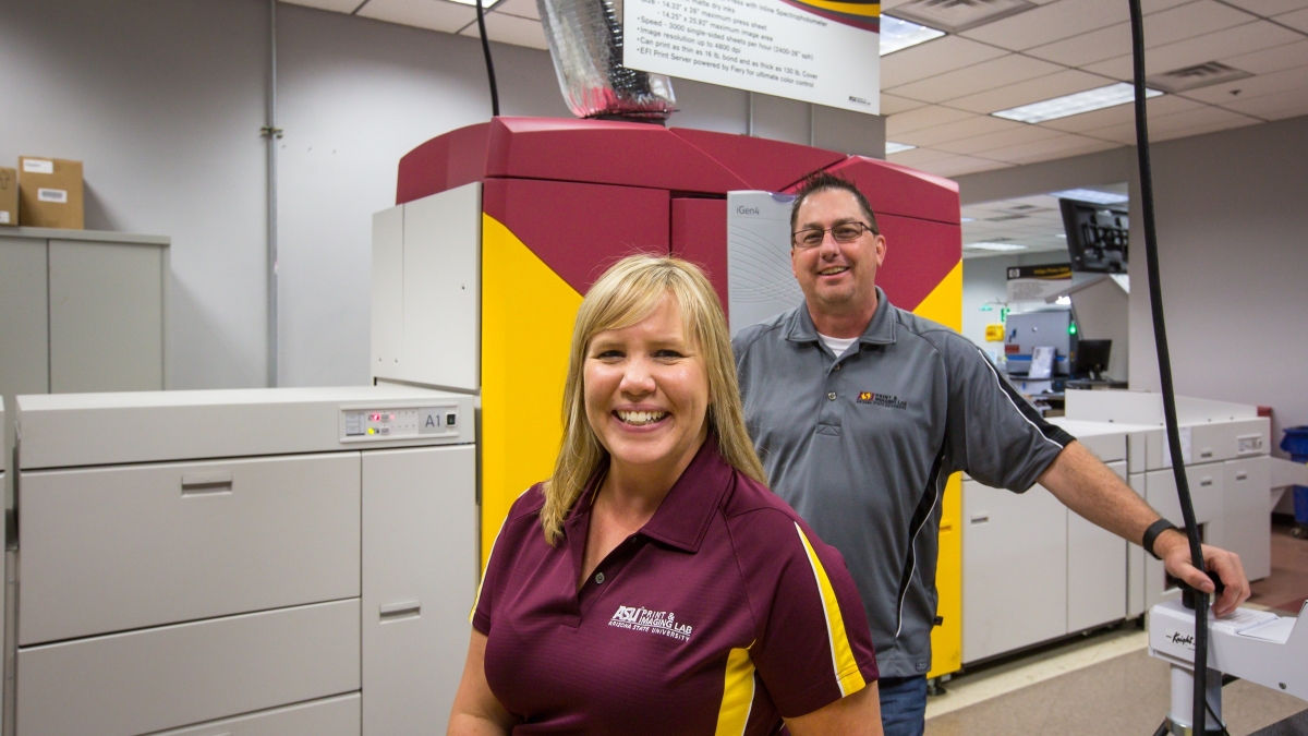 two ASU staff members posing in front of large printer