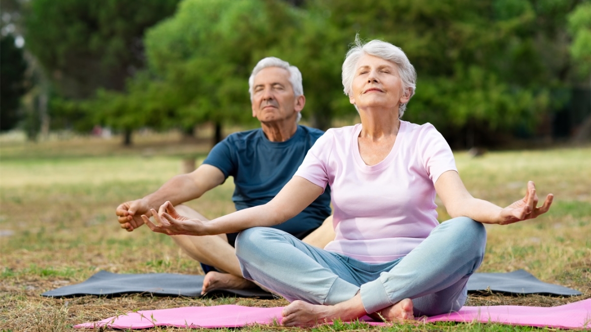 An older adult male and an older adult female meditating while seated on yoga mats in a field.