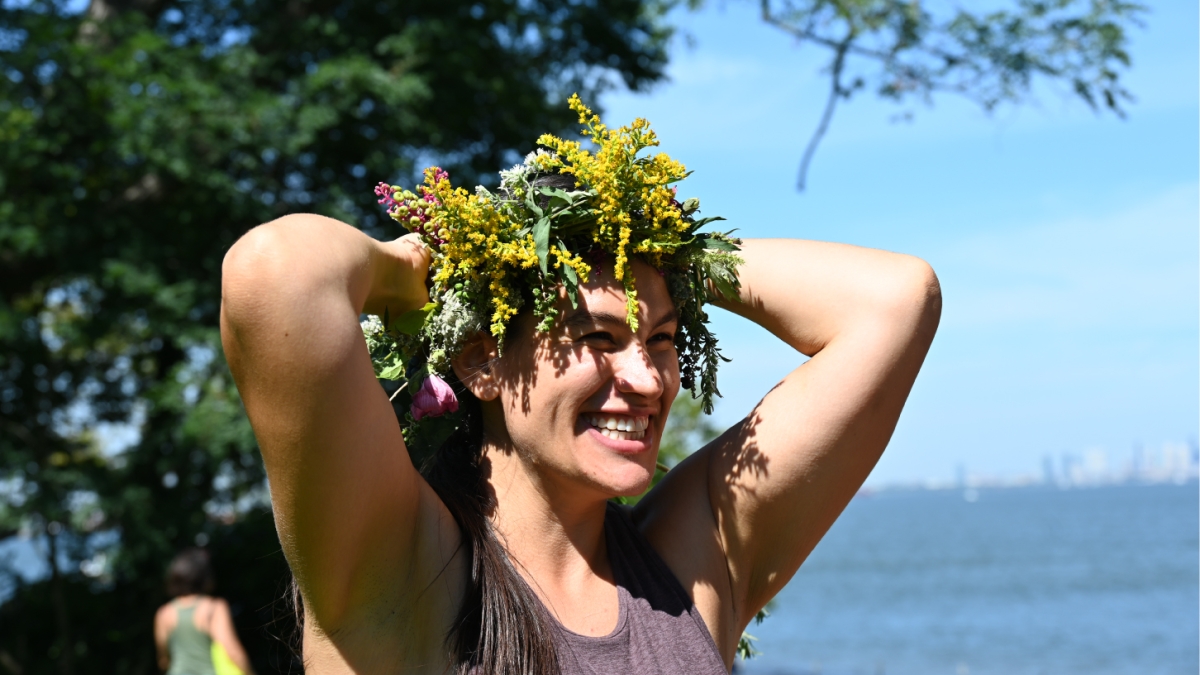 Aly Stoffo wearing a crown of greenery and smiling with her arms raised and a body of water and trees in the background.