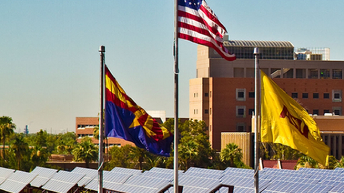Rooftop solar panels on the ASU Tempe campus are in the background of a three flags: The Arizona state flag, the United States flag and the ASU flag.