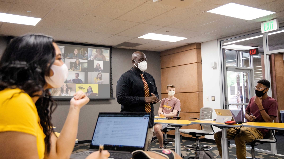 A teacher wearing a mask speaks to a socially distanced classroom of masked college students with a Zoom screen behind him of more students