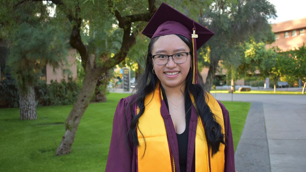 Fall 2020 graduate Monica Orillo poses in her maroon and gold graduation regalia, including a gown, stole, and mortarboard. A tree and grassy lawn are in the background.