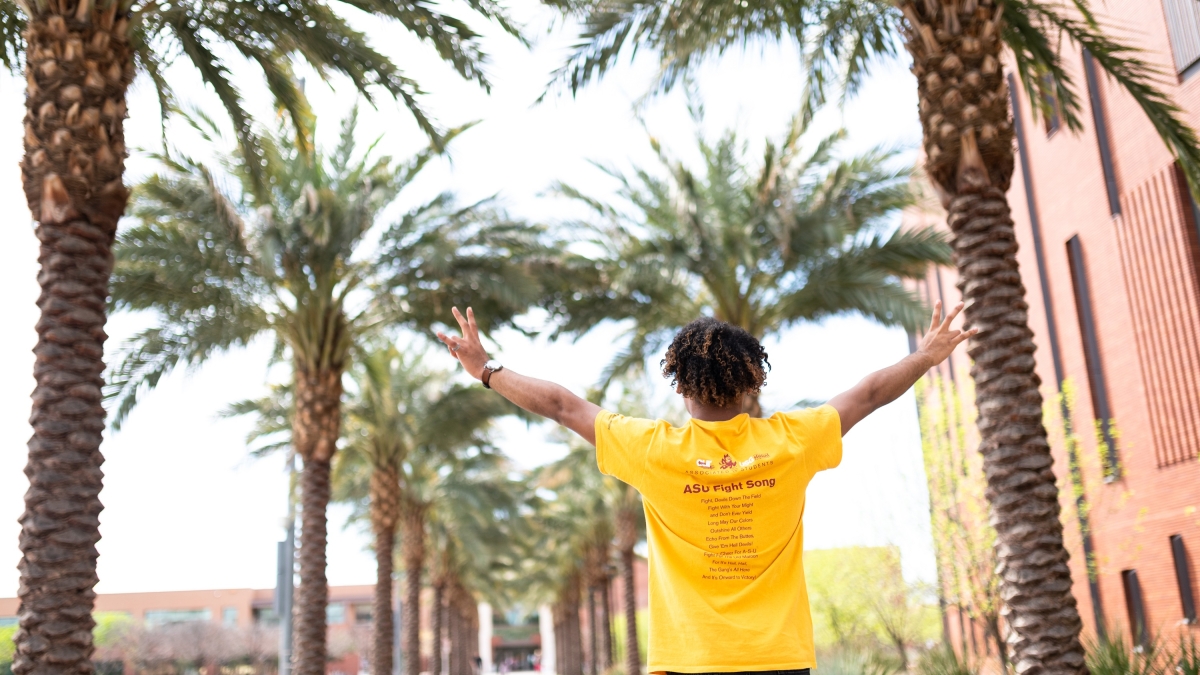 Student in a gold shirt on palm walk with his hands outstretched
