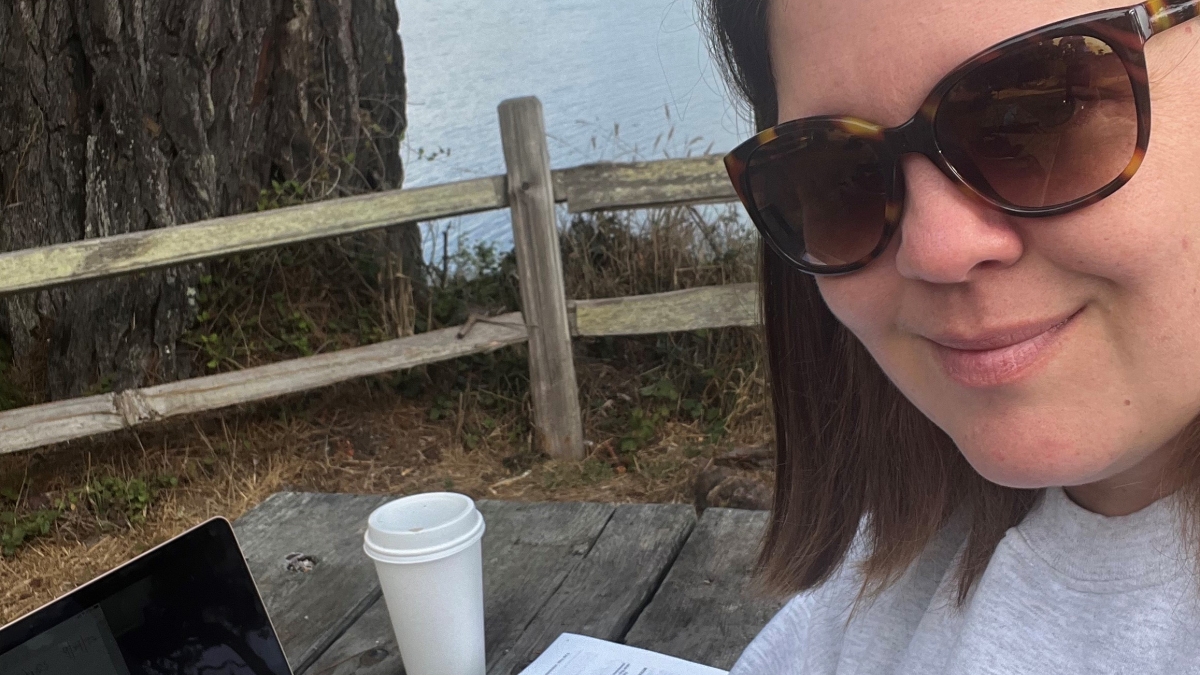 Portrait of ASU Online student Shawna Brechbill wearing sunglasses and sitting by the ocean with her laptop, some paper and a coffee.