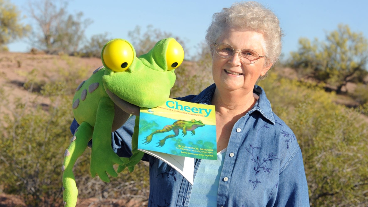 Betty Davidson holding a studded-animal frog with her children's book in its mouth, with an outdoor setting behind her.