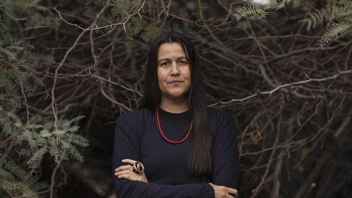 ASU Pulitzer Prize-winner Natalie Diaz wears a black shirt and red and black beaded necklaces as she stands with arms crossed against a backdrop of foliage.