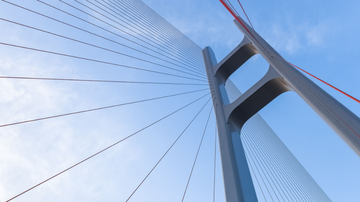 A gray suspension bridge is viewed from below with a blue sky behind it.
