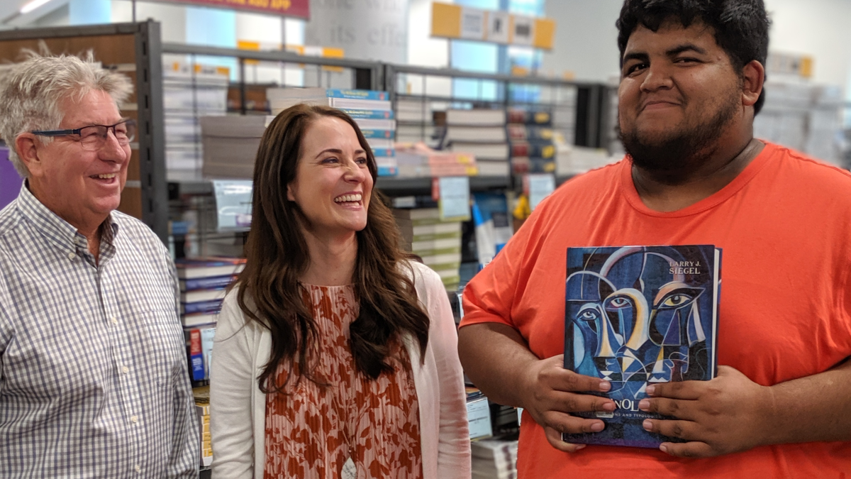 Representatives from ASU Bookstores and Follett (man and woman) smile with student Allan Valles Sanchez while he holds up a criminology and criminal justice textbook